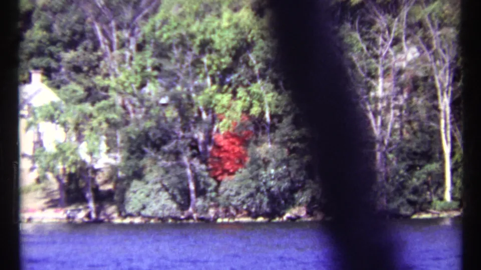 1967 Toronto home-movie still showing a wooded shoreline, pale house, red object in foliage, and blue water below.