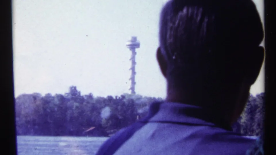 1967 Toronto home-movie frame of a silhouetted adult man facing water with a distant tower-like structure on the far shore.