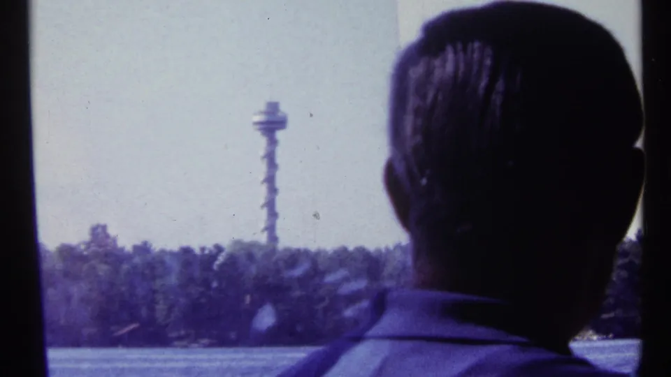 8mm home-movie view of a passenger's head and shoulders against a boat window, with water, trees, and a distant tower-like structure outside.