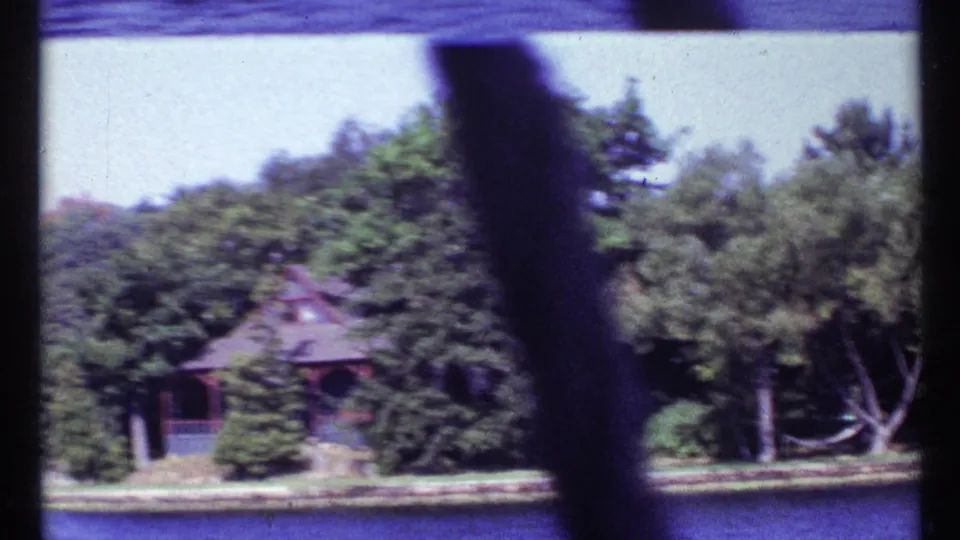 The boat passes a wooded waterfront with a small house or boathouse visible through the window frame in 1960s Canadian home-movie footage.