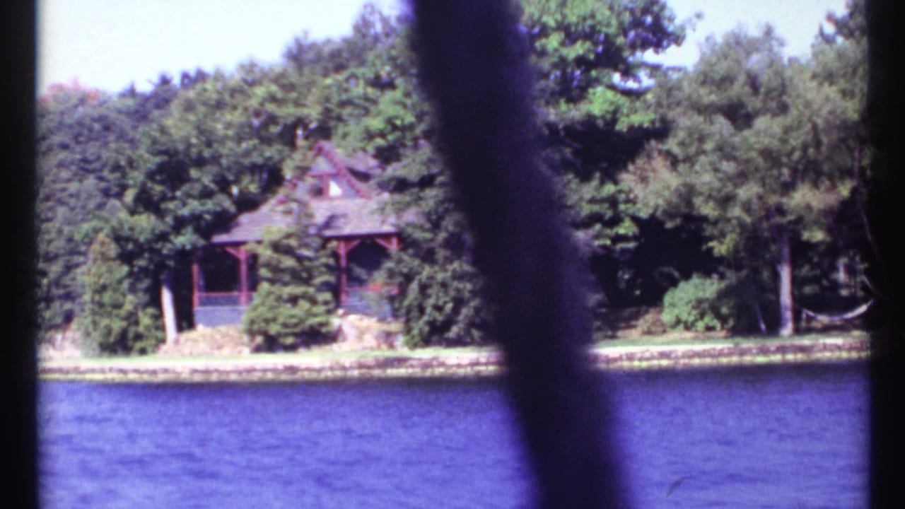 Cabin on a wooded shoreline across blue lake water, partly blocked by dark vertical foreground shapes.