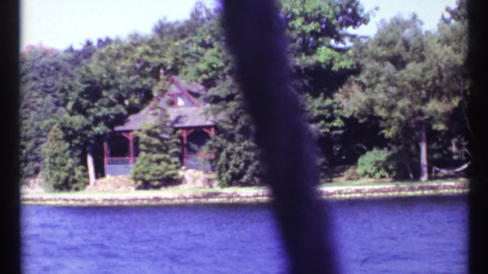 1967 8mm frame from a moving boat showing a rustic cabin among trees on a lake shore near Toronto, Canada, with foreground obstruction.