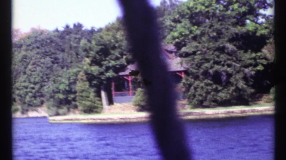 Soft 1967 8mm home-movie frame showing a cabin partly hidden by trees on a wooded lake shore, filmed from a boat near Toronto, Canada.