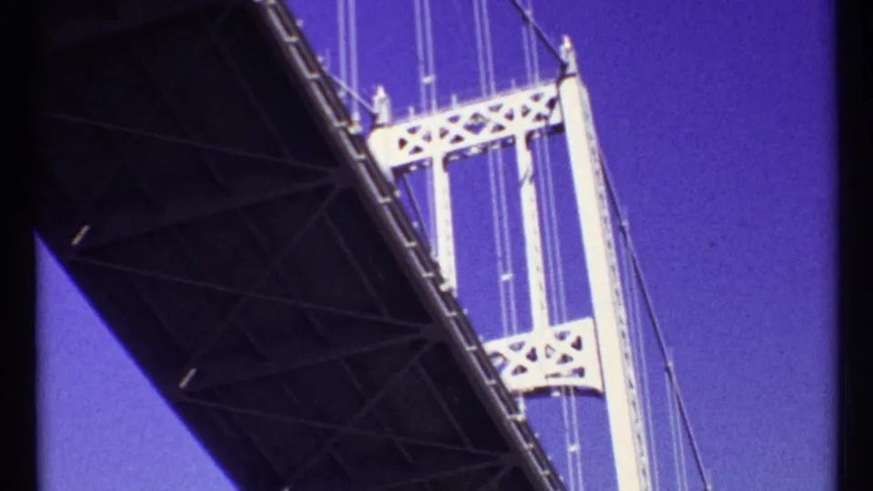 1967 Toronto home-movie frame looking upward from a boat beneath a bridge, showing its underside, tower, cables, and blue sky.