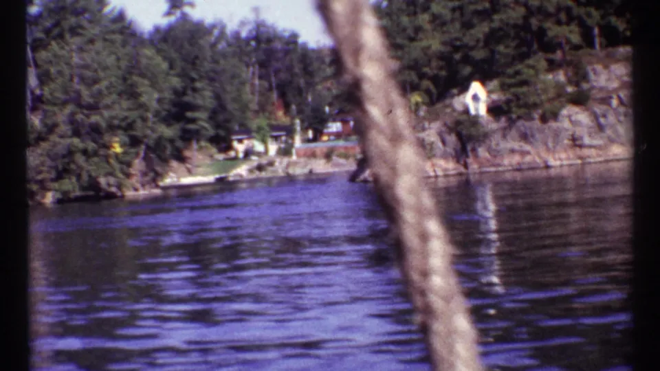 1967 Toronto 8mm frame looking across water toward a wooded rocky shore with small buildings visible.