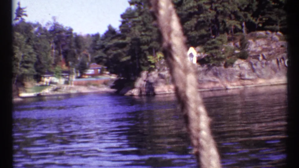1967 Toronto 8mm frame showing water, wooded shoreline, rock outcrops, and small structures from a handheld vantage point.