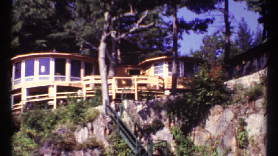1967 Toronto home-movie view of a lakeside modernist house with curved wood balcony, large windows, trees, and rock ledge.