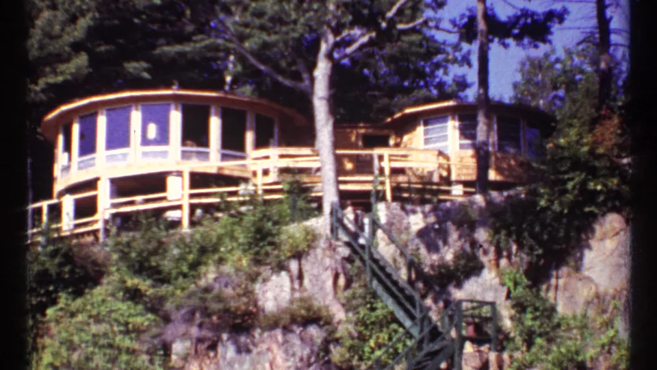 1967 Toronto home-movie frame with the curved lakeside house, wooden deck railings, exterior steps, tree trunks, and cliffside vegetation.