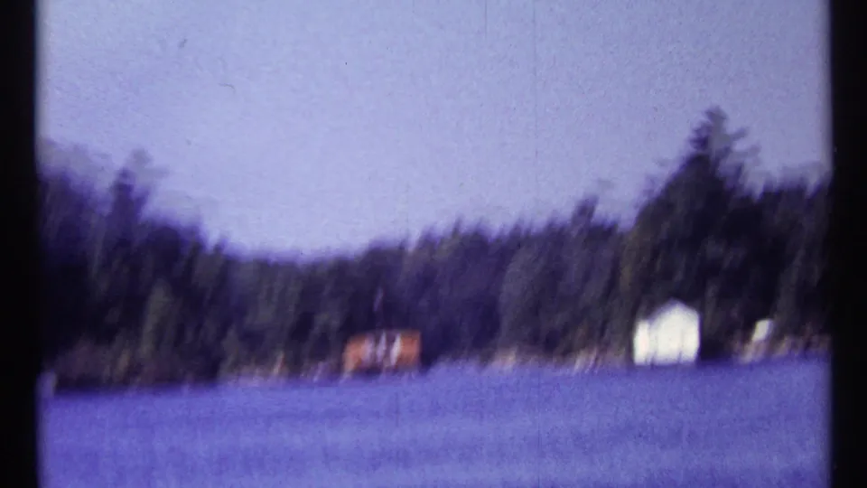 Soft 8mm frame from 1967 Toronto footage showing blue lake water, distant wooded shoreline, and small lakeside buildings.