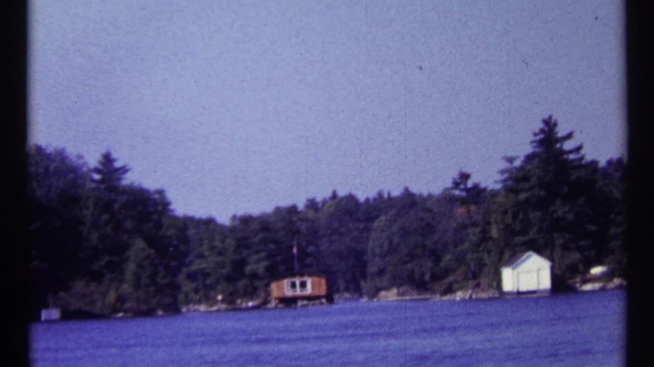 1967 home-movie frame of a wooded lakeside shoreline in the Toronto, Canada metadata location, with small shore buildings across calm water.