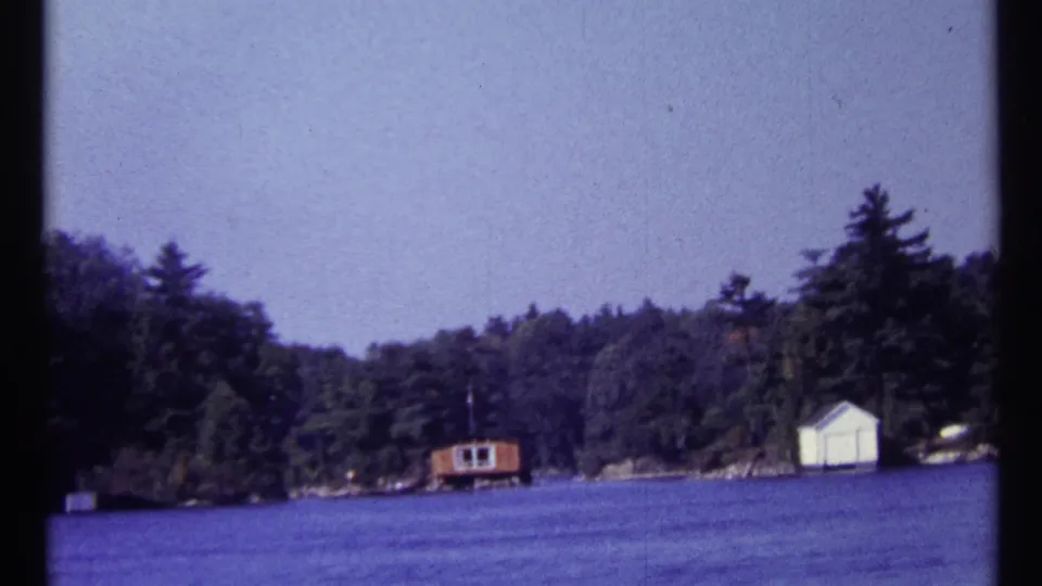 1967 home-movie frame of a wooded lakeside shoreline in the Toronto, Canada metadata location, with small shore buildings across calm water.