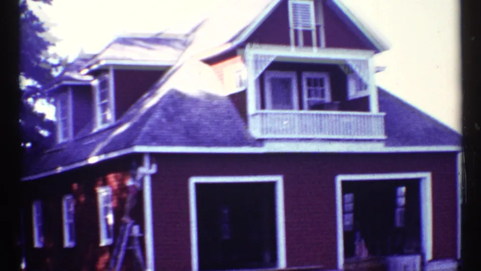 Red lakeside house exterior in 1967 home-movie footage, showing white trim, dormers, balcony railing, open lower bays, and a ladder near the wall.