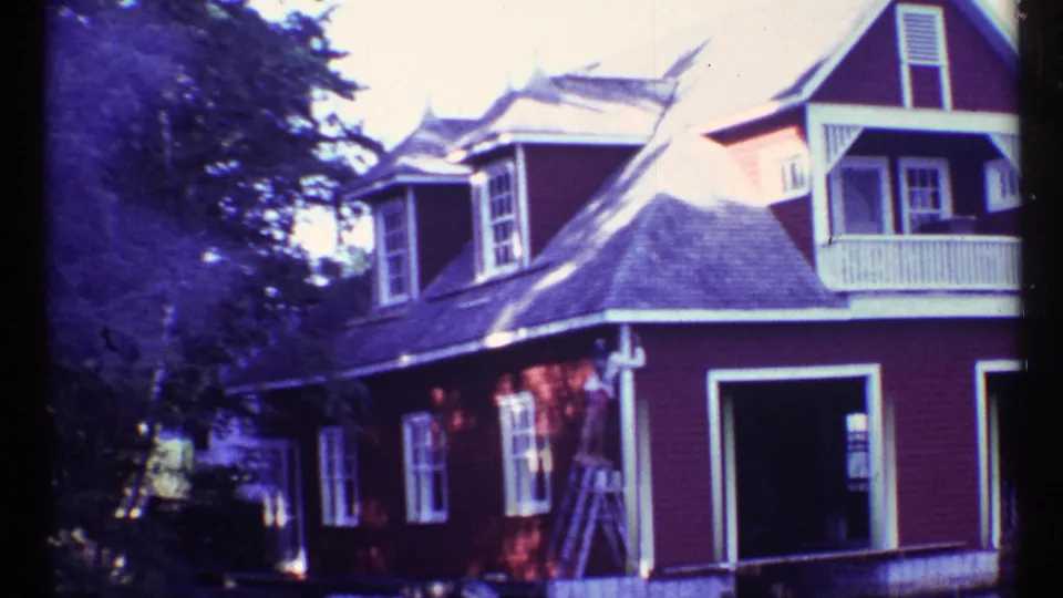 1967 home-movie frame of a red house exterior with white trim and ladder, where a person appears to be standing near the wall.