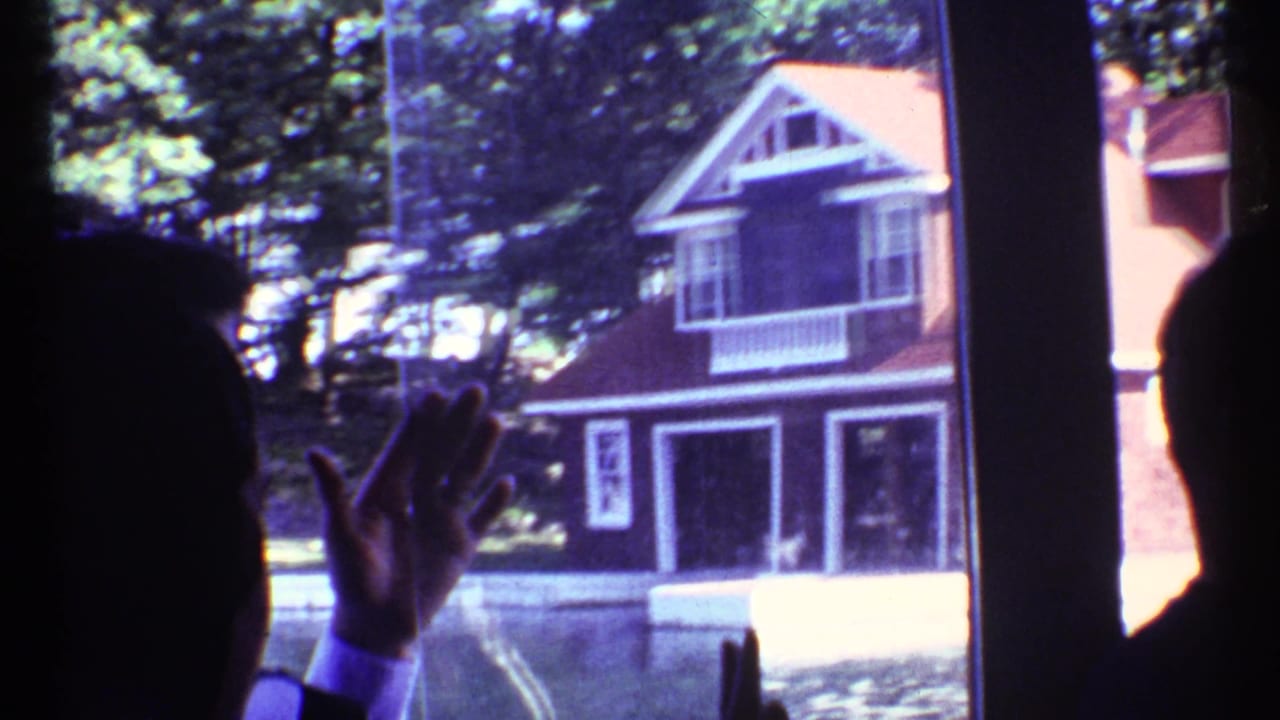 Silhouetted person raises a hand near a window looking toward a red house outside.