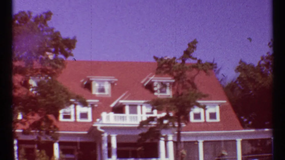 1967 Toronto frame of a red-roofed residential house with dormers, white columns, balcony railing, and trees in front.