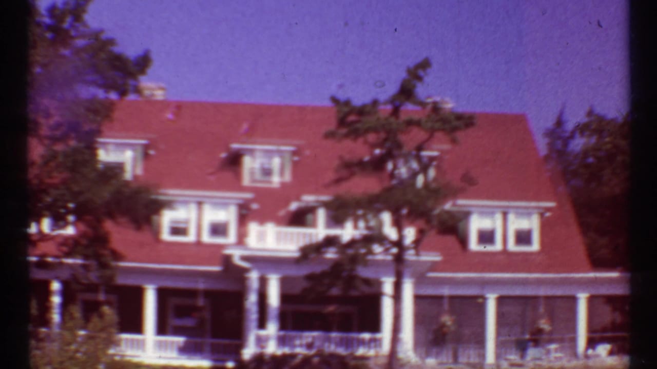 Large red-roofed lakeside house with white porch columns and trees in front.