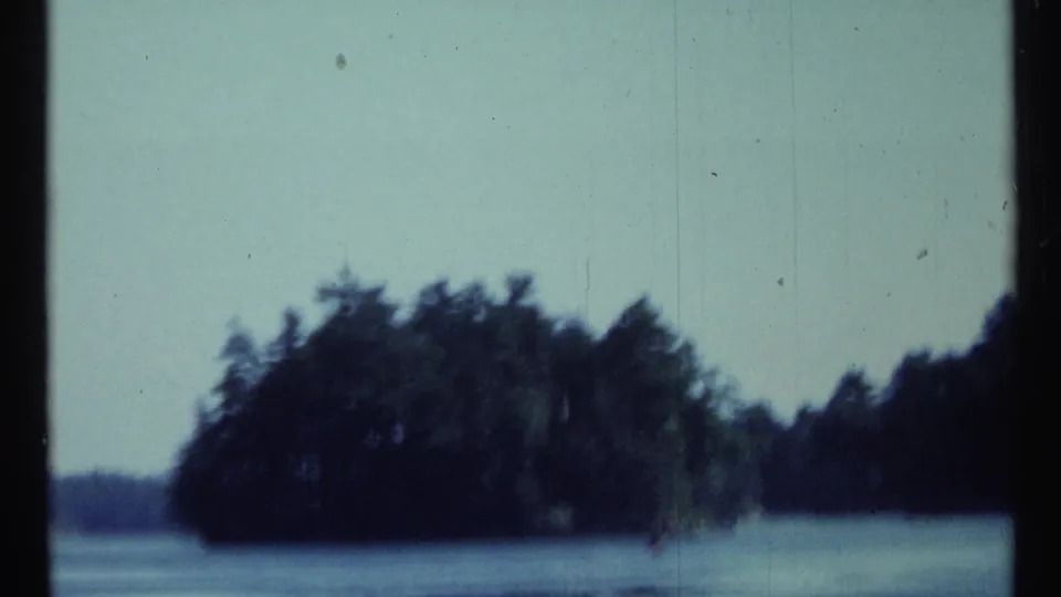 1967 Toronto boat-view frame showing a small wooded island or shoreline mass across calm water, with soft focus and visible film grain.