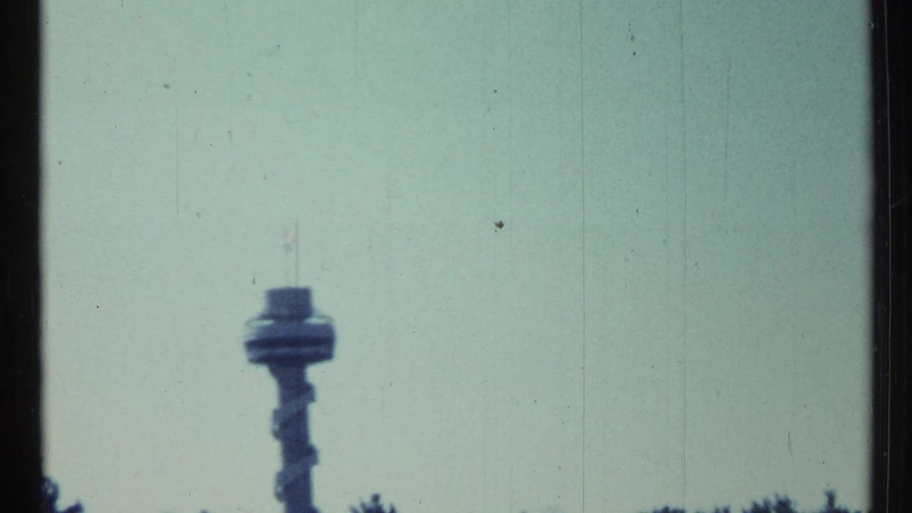 Toronto-area 1967 home-movie frame with a distant airport control tower visible above the treeline in soft focus.