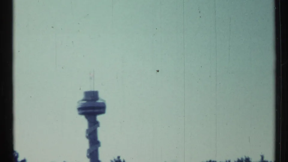 Toronto-area 1967 home-movie frame with a distant airport control tower visible above the treeline in soft focus.