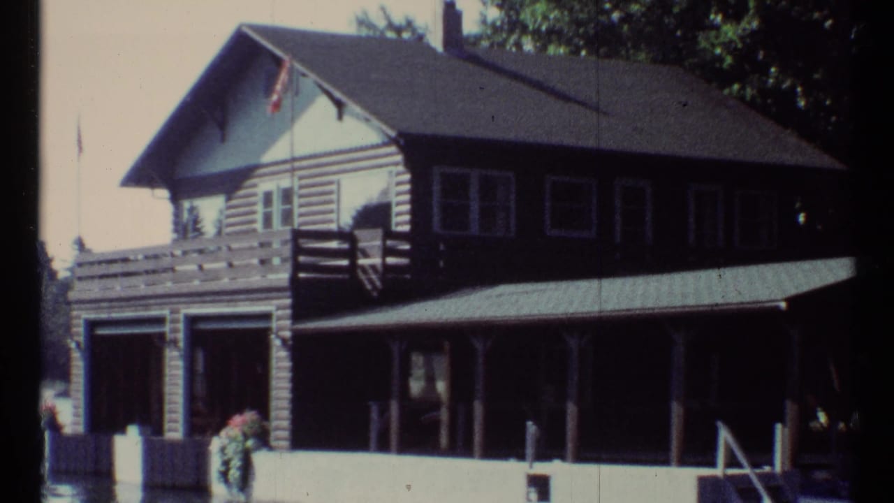 Two-story lakeside log cabin with porch, boat garage, dock pilings, flag, trees, and water.