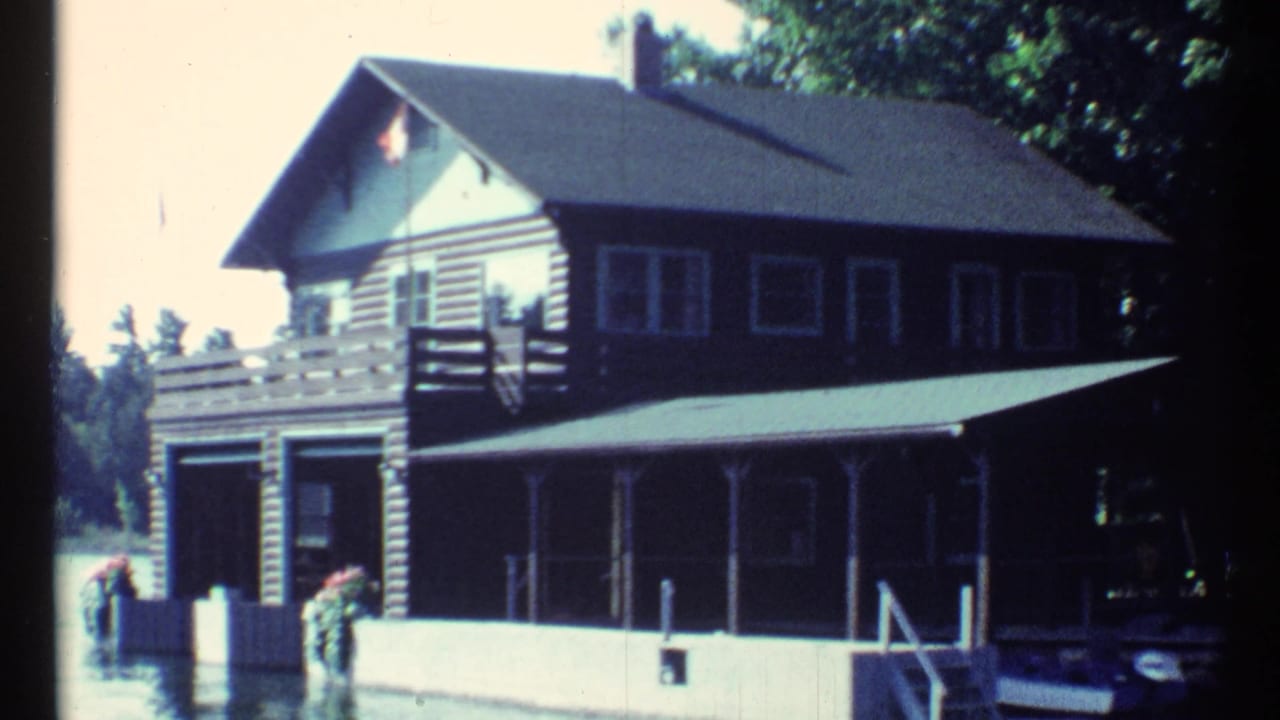 Lakeside cabin exterior with boat garage doors, porch, dock pilings, trees, water, and small figures near the dock.