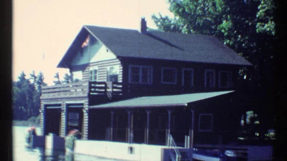 1967 Toronto home-movie frame of a lakeside log cabin with a porch, boat garage, Canadian flag, dock pilings, trees, and rippled water.