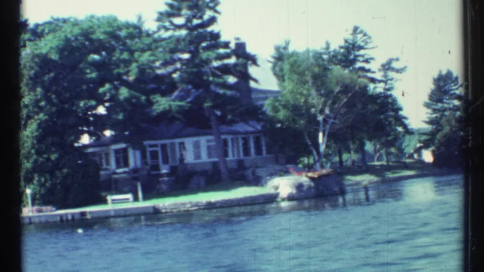Wider 1967 Toronto lake view showing a waterfront house partly obscured by trees, a dock edge, lawn, rocks, and calm water in the foreground.