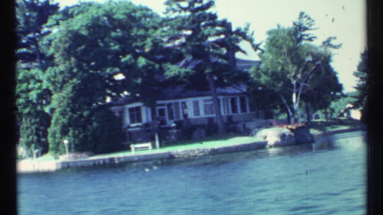 Large lakeside house partly hidden by trees, seen across water from a moving boat.