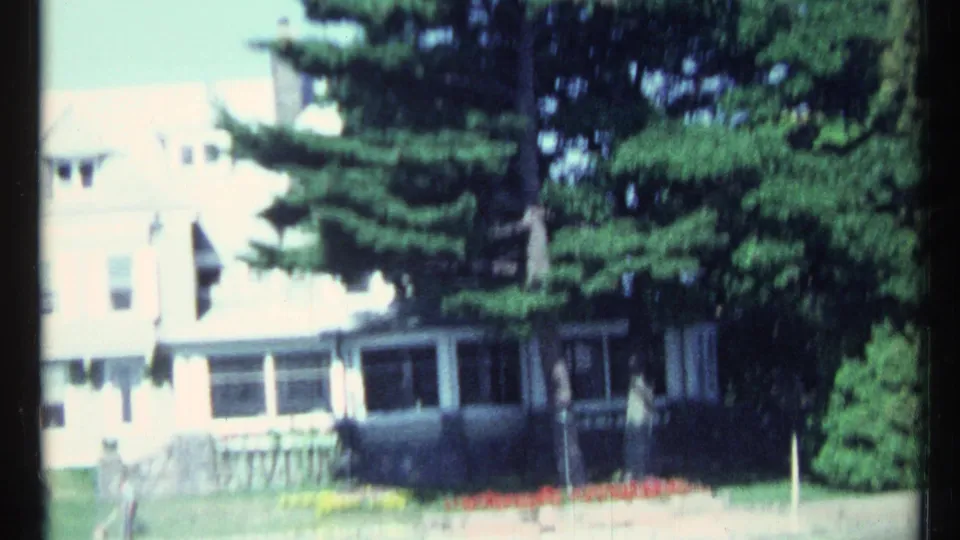 1967 8mm frame showing a light-colored lakeside house with porch windows, lawn plantings, stonework, and large evergreens.