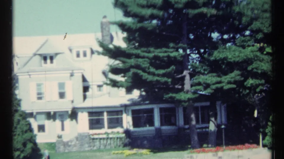 End frame of the 1967 Toronto lakeside house view, showing the residence, stone base, porch area, lawn, flowers, and tall evergreens.