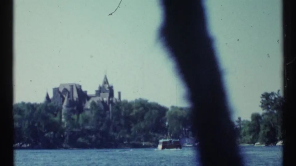 1967 Toronto home-movie frame showing water, a small boat, and a distant turreted building beyond a tree-lined shore.