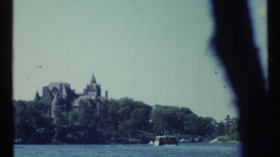 1967 Toronto-area home-movie view across water toward a wooded shoreline and distant castle-like mansion, partly obscured by dark foreground shapes.
