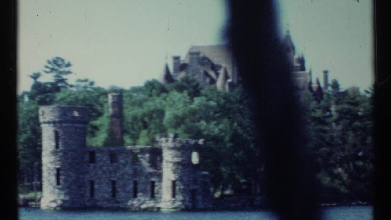 Stone ruin beside wooded shoreline with a large mansion rising behind trees.