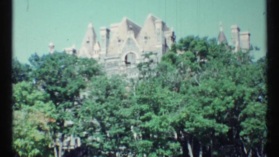 1967 Toronto home-movie frame showing turreted stone architecture above a thick line of summer trees.
