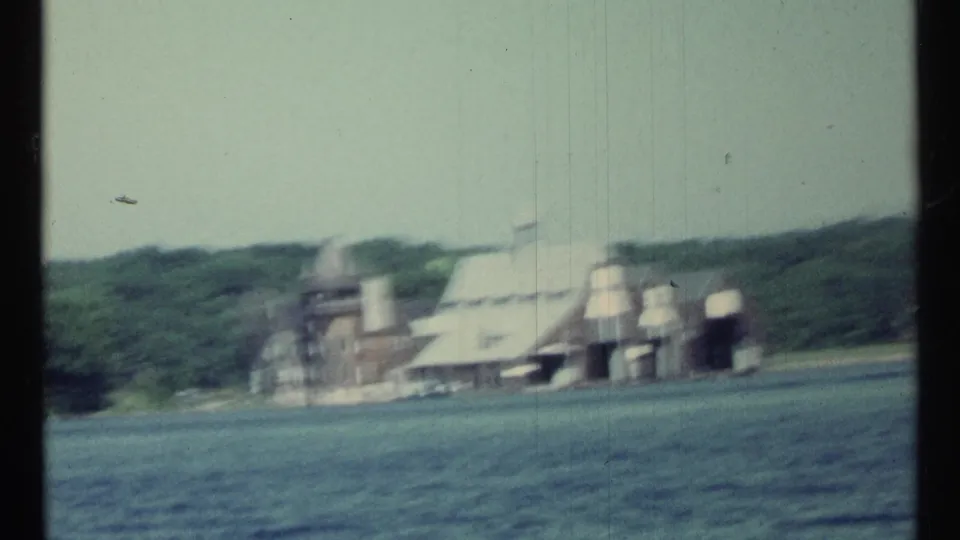 1967 8mm frame from near Toronto showing a distant estate-style building on a wooded lakeshore viewed from a boat.