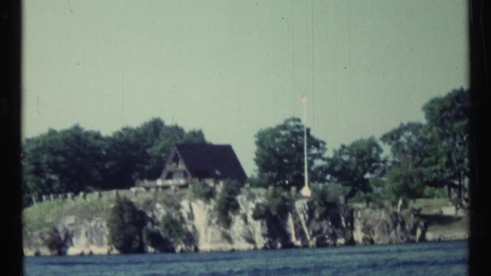 Later frame from the 1967 Toronto-area lake view showing rocky shore, trees, a small roofed structure, and a flagpole.