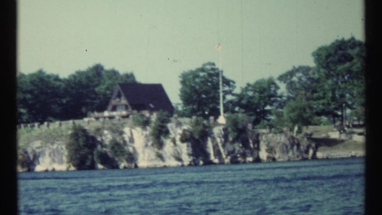 A-frame cabin on a rocky wooded shoreline seen across water.