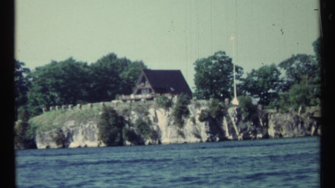 Rocky lakeshore with cabin, trees, and flagpole viewed from a boat.