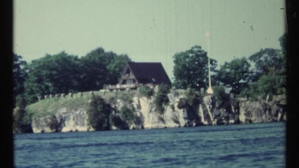 1967 Canada 8mm frame showing a cabin and flagpole on a wooded rocky shoreline from a moving boat perspective.