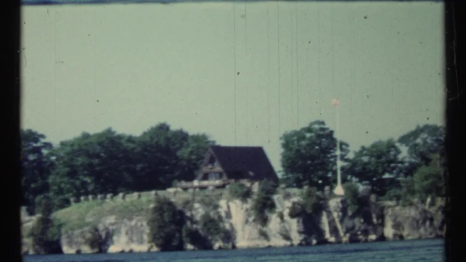 1967 Canada frame of a quiet rocky shoreline with an A-frame cabin, trees, flagpole, and water in the foreground.