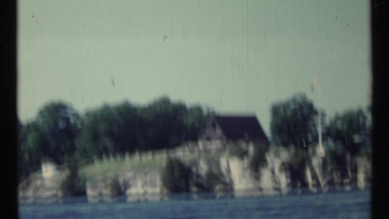 Boat-level view of rocky shoreline, trees, water, and a distant building.