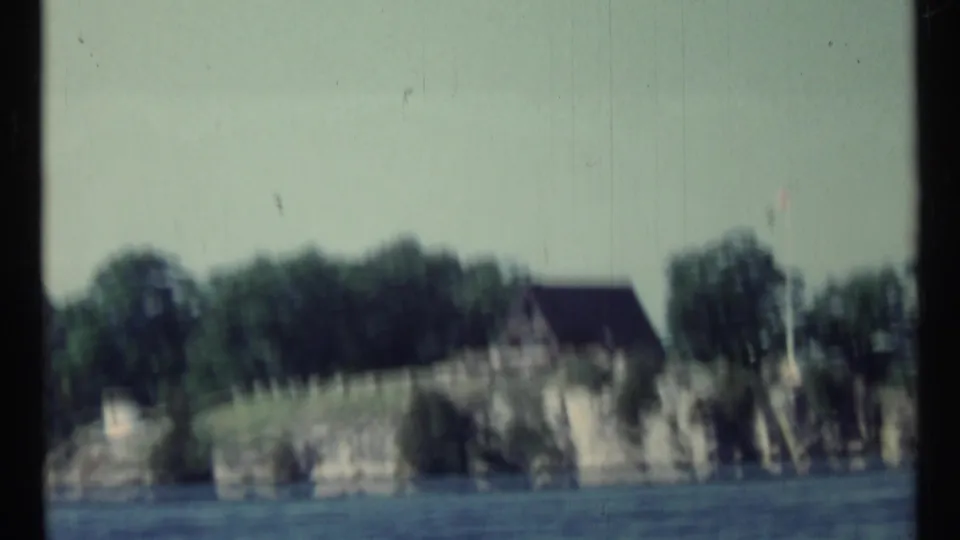 1967 Toronto-area 8mm frame showing a shoreline from the water, with rocky banks, trees, and a distant building in soft focus.