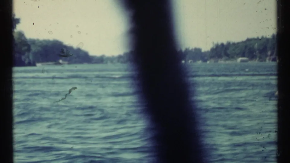 1967 Toronto-area 8mm frame looking across rippling water toward a wooded shoreline, partly blocked by a dark vertical element.