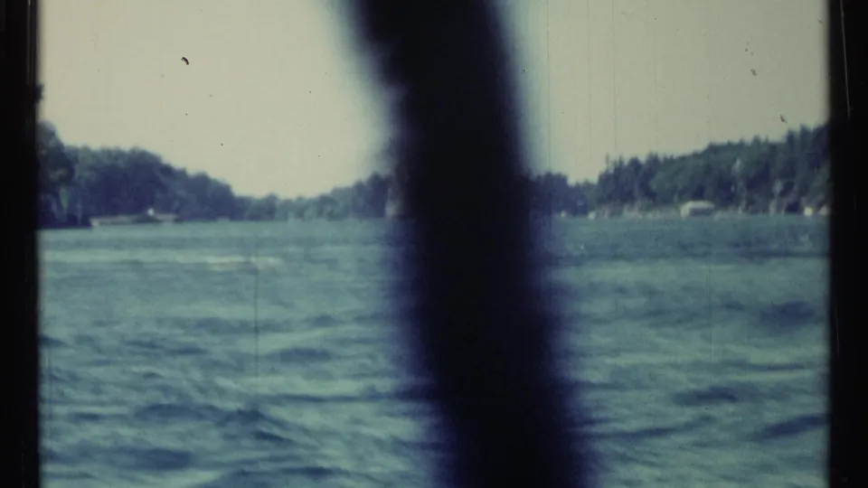 1967 Toronto-area 8mm frame of blue-green water and distant wooded banks, with a dark foreground element cutting through the view.