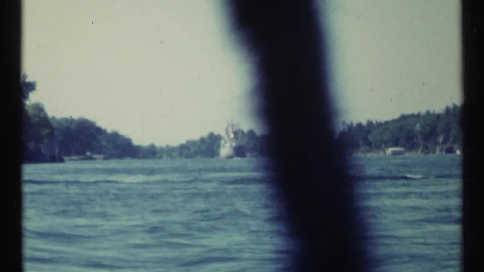 1967 Toronto-area 8mm frame showing rippling water, wooded shoreline, and a dark vertical obstruction from a boat-level viewpoint.