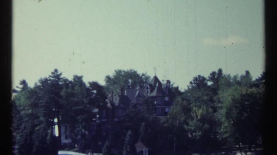 1967 home-movie frame of a wooded Toronto waterfront with a large mansion and smaller structure near the shore.