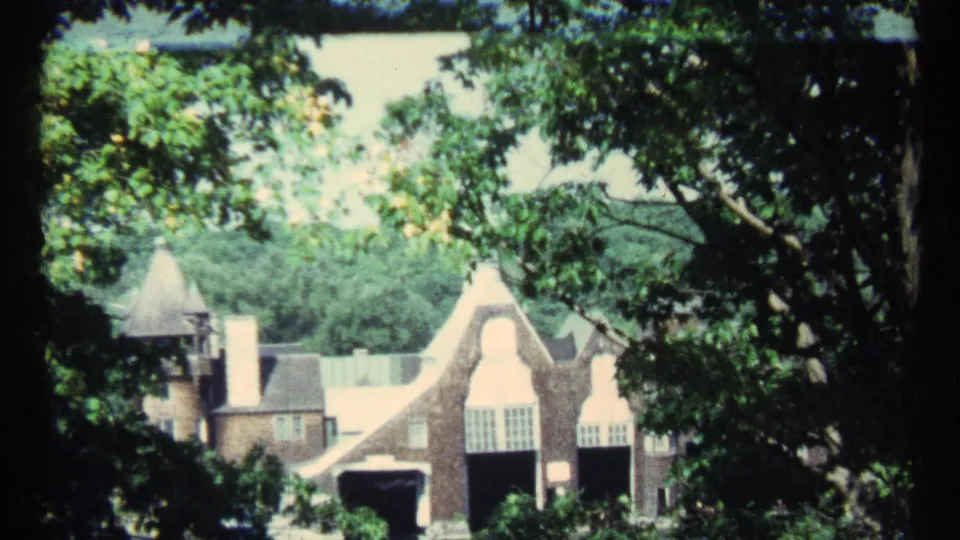 1967 Toronto home-movie frame of the estate viewed through trees, showing Tudor-style gables, chimneys, windows, and deep arched openings.