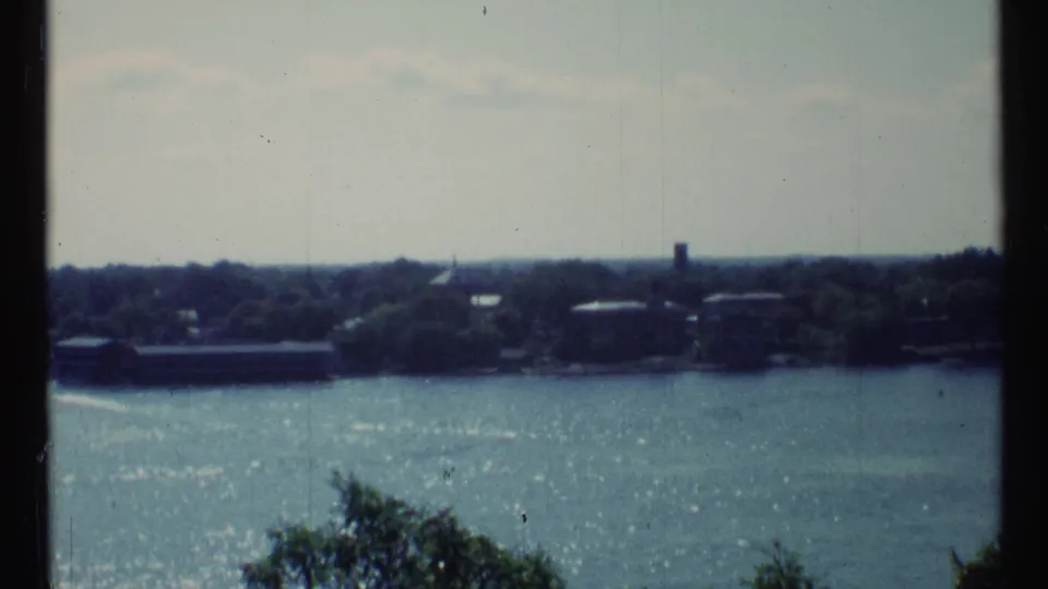 Soft 1967 Toronto river scene with shimmering water, wooded shoreline, and low structures in the distance.