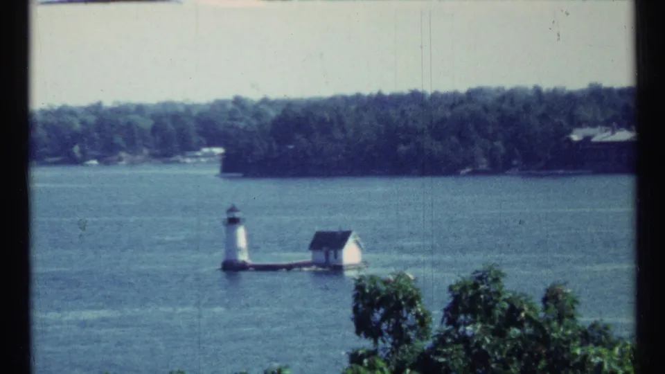 Soft 8mm frame of a small lighthouse and white building on a lake near Toronto, with foreground trees and a wooded shoreline.