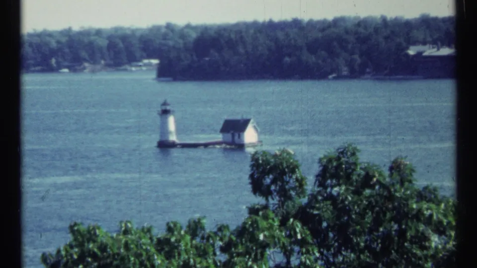1967 home-movie frame of a quiet lake scene near Toronto, centered on a small lighthouse and white building beside leafy trees.
